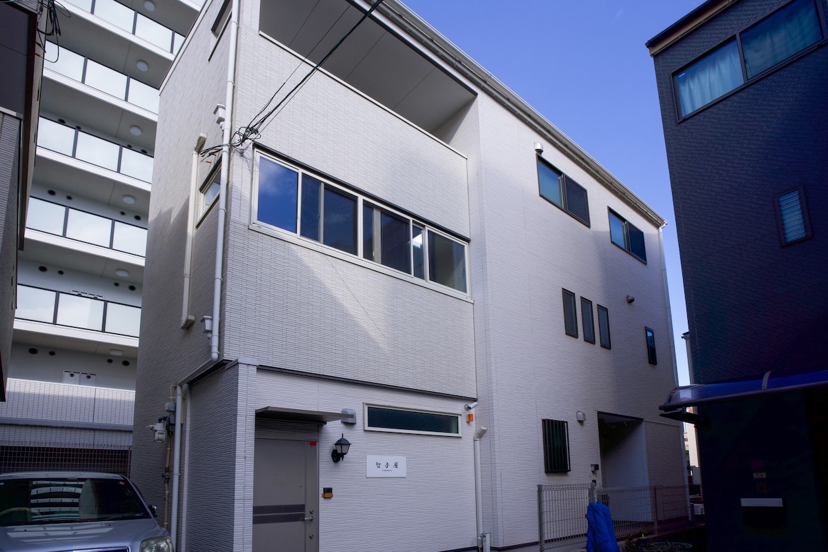 The three-story villa features a light-colored exterior with large windows, providing ample natural light. A driveway with a parked vehicle is visible, alongside a pathway leading to the entrance. The building is situated in a tranquil setting, surrounded by neighboring structures.