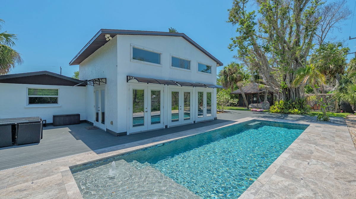 A spacious house is shown with large glass windows, allowing natural light to fill the interior. The solar heated pool glimmers in the foreground, surrounded by a well-maintained patio area. Lush greenery provides a natural backdrop, enhancing the privacy of the outdoor space.