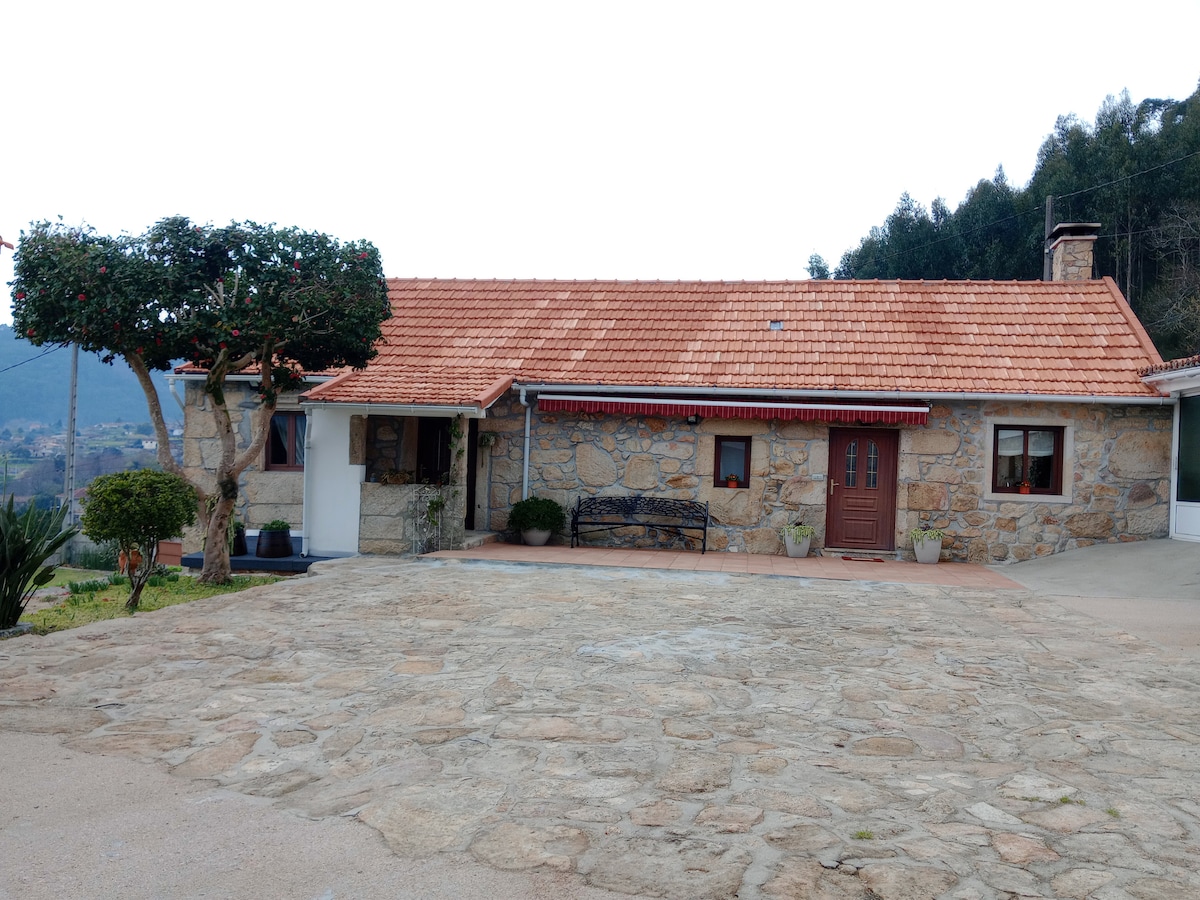 A charming stone house is shown with a red-tiled roof and a front patio area. A wooden door is centrally positioned, flanked by plants. The surrounding landscape features greenery and hills in the background, enhancing the rural setting.