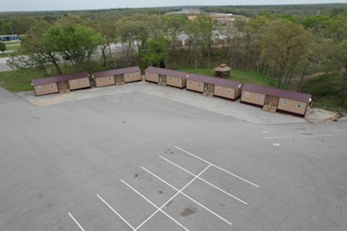 Aerial view of a row of duplex-style cabins situated in a gravel outdoor area. Each cabin features a uniform design with a pitched roof and wooden doors. Surrounding trees provide a natural backdrop, creating a sense of space between the cabins and the parking area.