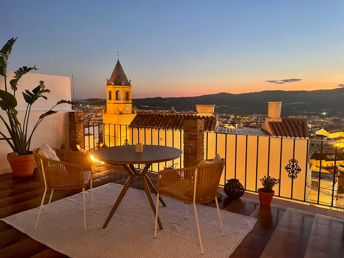 A spacious terrace offers a round table with two woven chairs, set against a backdrop of the evening sky transitioning from orange to deep blue. The image features a plant in a pot, providing a touch of greenery alongside views of the city and the distant mountains.