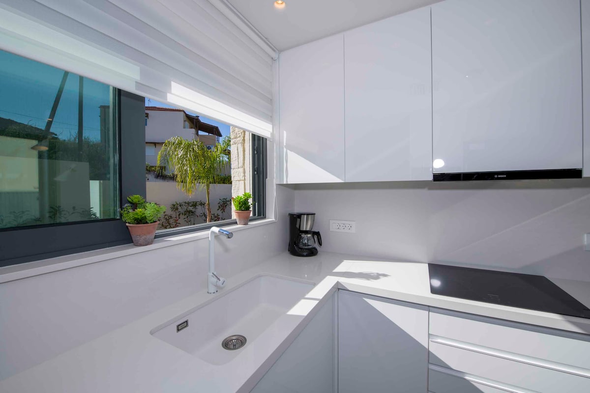 A modern kitchen space features sleek white cabinetry and a minimalist design. Natural light floods the area through a large window, accented by potted plants. A coffee maker and a stovetop are visible, contributing to the kitchen's functional layout.