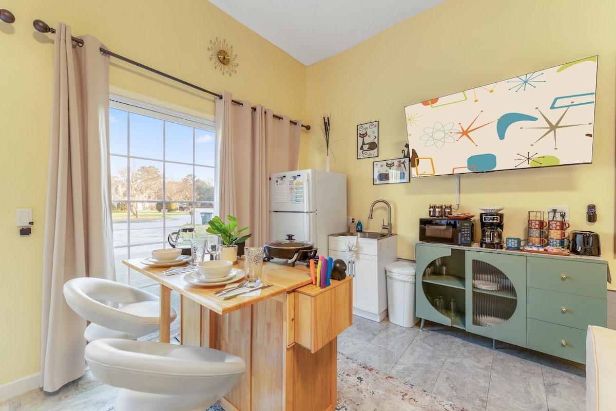 A well-appointed kitchenette is displayed, featuring a wooden table set for dining. Two modern chairs face a colorful 55" Smart TV mounted on the wall. A refrigerator, sink, and multiple kitchen appliances are visible, alongside ample natural light streaming through a large window.