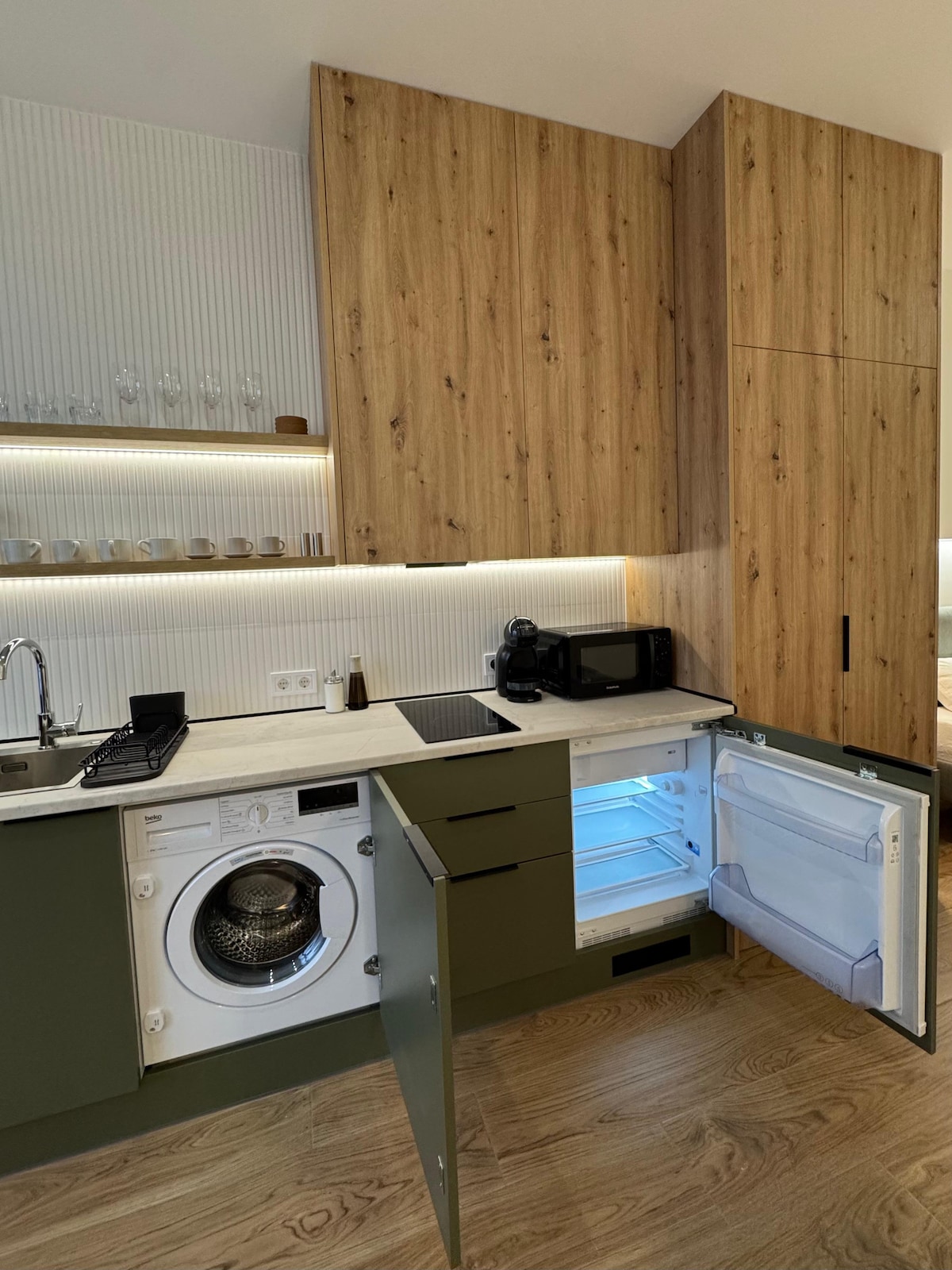 A modern kitchen is shown featuring a blend of wood and green cabinetry. A washing machine is visible beneath the counter, and an open refrigerator reveals bright interior lighting. Essential appliances, including a microwave and coffee maker, are also present, enhancing the functionality of the space.