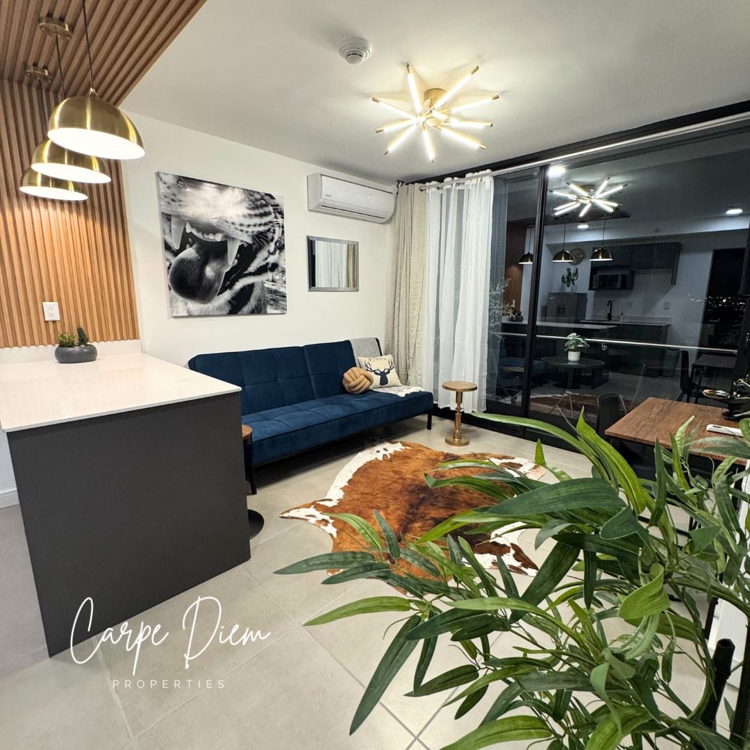 A modern living area showcases a comfortable dark blue sofa and a light wood dining table. The space features stylish pendant lights and a large window that offers views of the city. A decorative rug adds warmth to the neutral-toned floor.