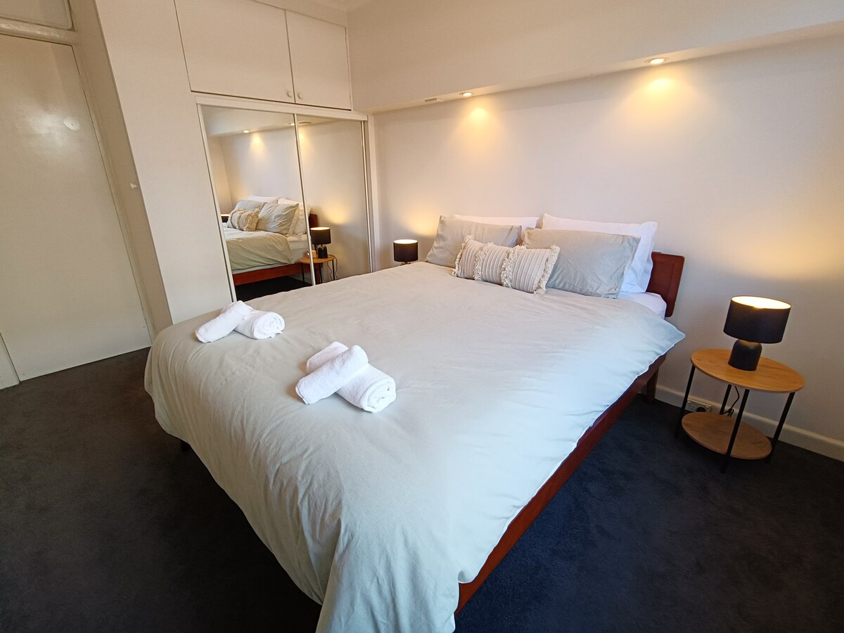 A well-lit bedroom features a spacious bed dressed in light bedding. Two towels are neatly placed at the foot of the bed. Soft, ambient lighting is provided by two bedside lamps. A wardrobe with mirrored doors reflects the room’s simplicity and comfort.