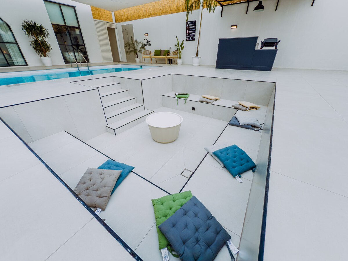 An outdoor seating area is integrated into the sleek pool design, featuring a sunken lounge with a circular table at the center. Several cushions in varied colors are positioned around the seating area, offering a comfortable spot for relaxation beside the inviting pool.