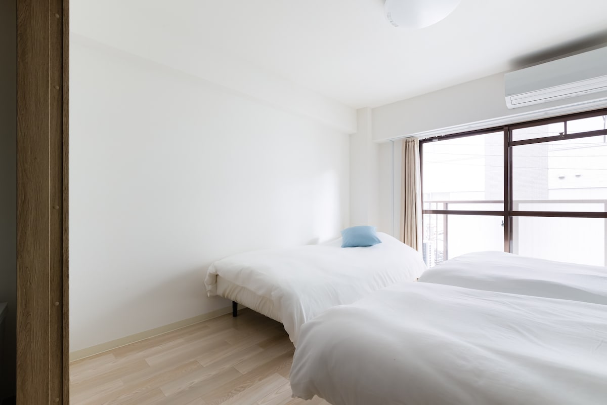 A bright sleeping area features two spacious beds, each with white bedding and a light blue accent pillow. Natural light enters through a large window, complemented by light curtains, contributing to a calm atmosphere. A neutral color palette enhances the room's simplicity.