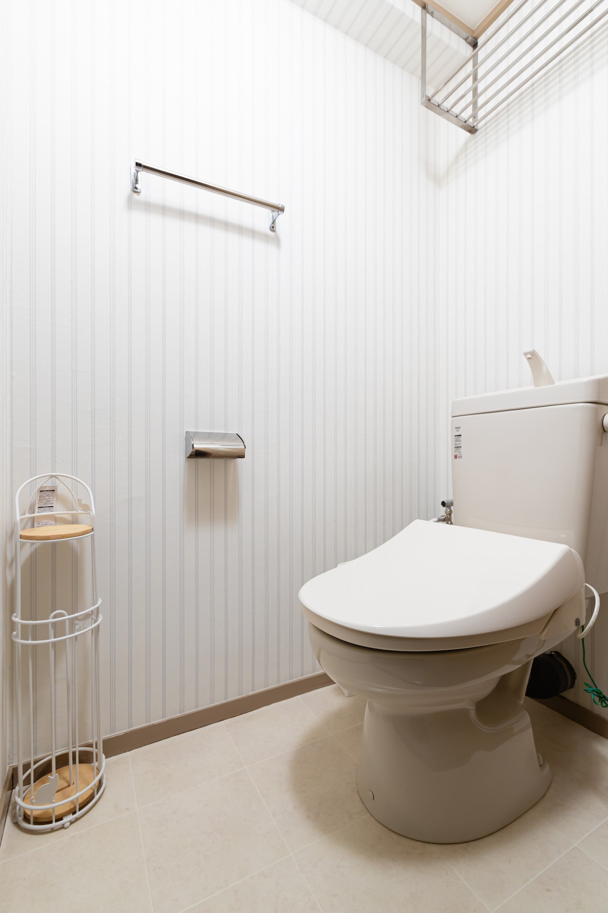 A clean and tidy bathroom features a modern toilet positioned on light-colored tiled flooring. Vertical striped wallpaper adds texture to the walls. A hand towel rack is mounted above the toilet, and a decorative toilet paper holder stands beside it.