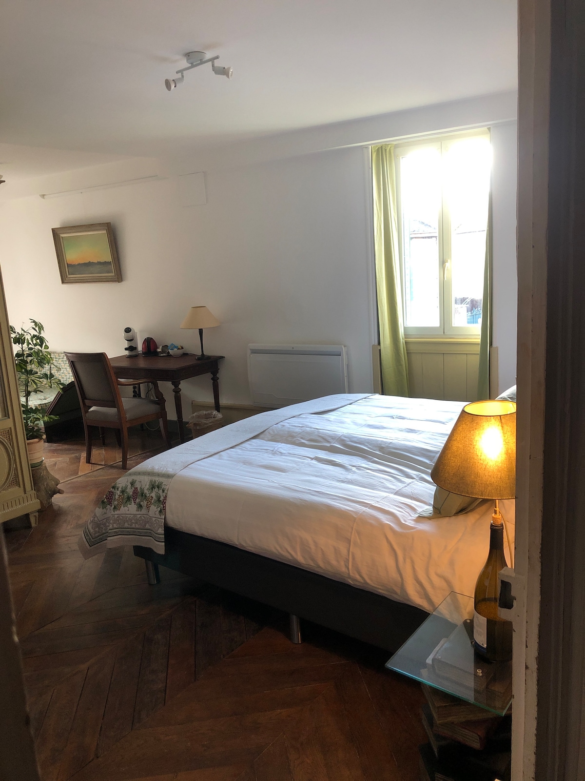 A spacious bedroom is illuminated by natural light from large windows draped with light-colored curtains. A comfortable bed is dressed in crisp white linens, complemented by a decorative throw. A small wooden desk and chair, along with a bedside lamp, add functionality to the inviting space.