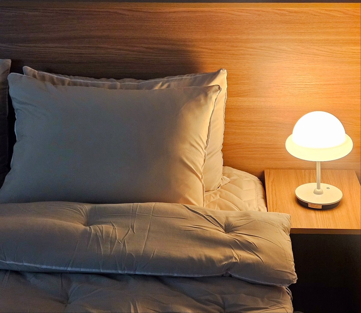 A spacious bed is dressed in soft, neutral bedding. A bedside table with a modern lamp is positioned next to the bed, softly illuminating the area. The warm wood headboard adds a touch of comfort to the sleeping space.