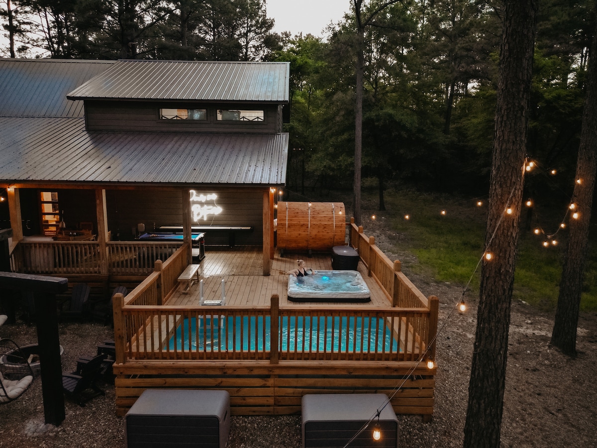 An outdoor deck features a hot tub by a wooden barrel sauna, both surrounded by a wooden railing. String lights provide a soft glow above the scene, while the adjacent pool area is visible. Tall trees create a natural backdrop, adding to the secluded feel of the space.
