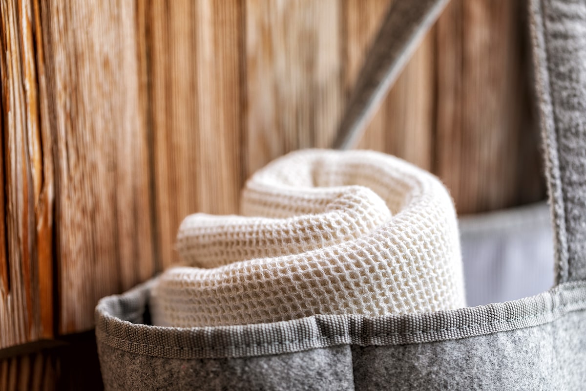 A neatly rolled white towel is placed inside a light gray felt storage basket. The basket is set against a backdrop of textured wooden panels, creating a calming and organized appearance.