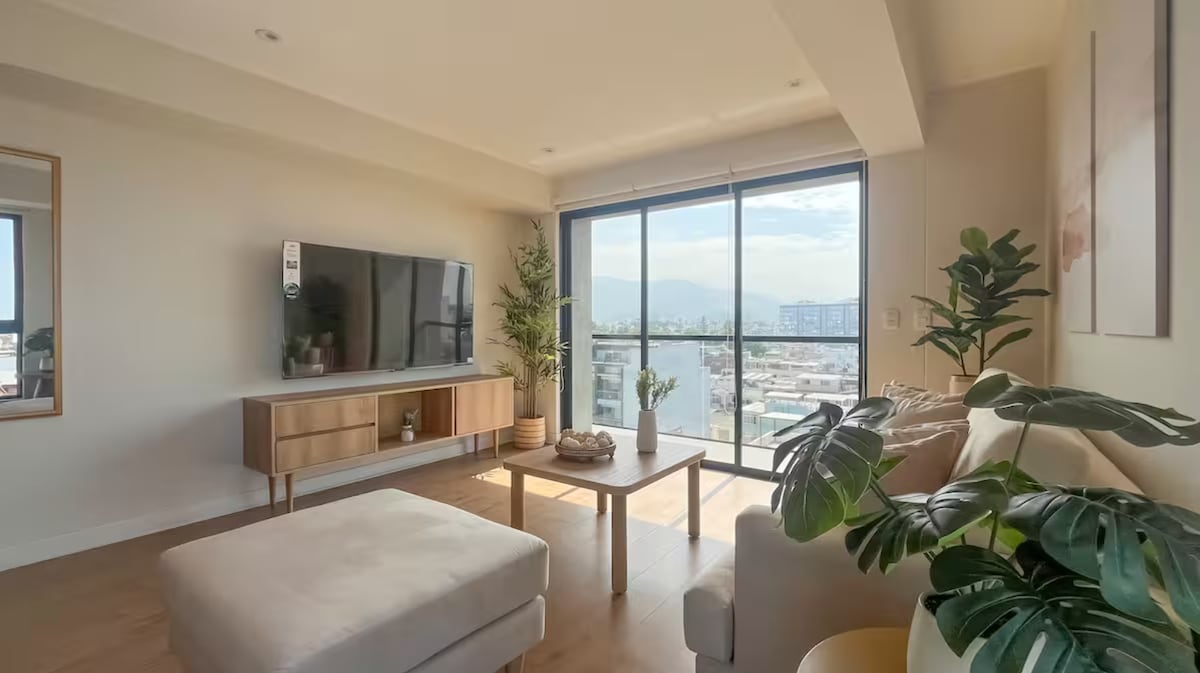 A bright and airy living area features a large window with panoramic city views. A smart TV is mounted on a stylish media console. A comfortable sofa and ottoman provide seating, accompanied by potted plants that add a touch of greenery to the space.