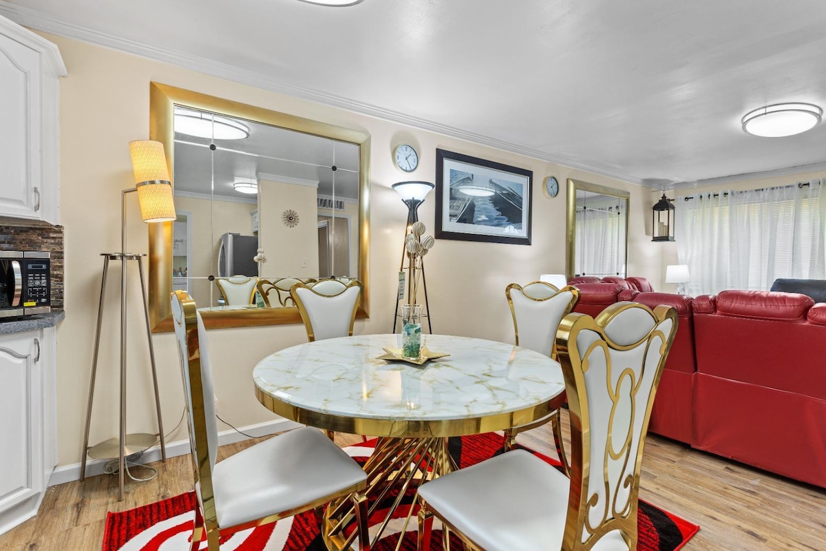 A dining area is depicted with a round marble table and four elegant chairs. A floor lamp and mirrors enhance the space. Nearby, a cozy seating area with red couches is visible, accompanied by light streaming through sheer curtains.