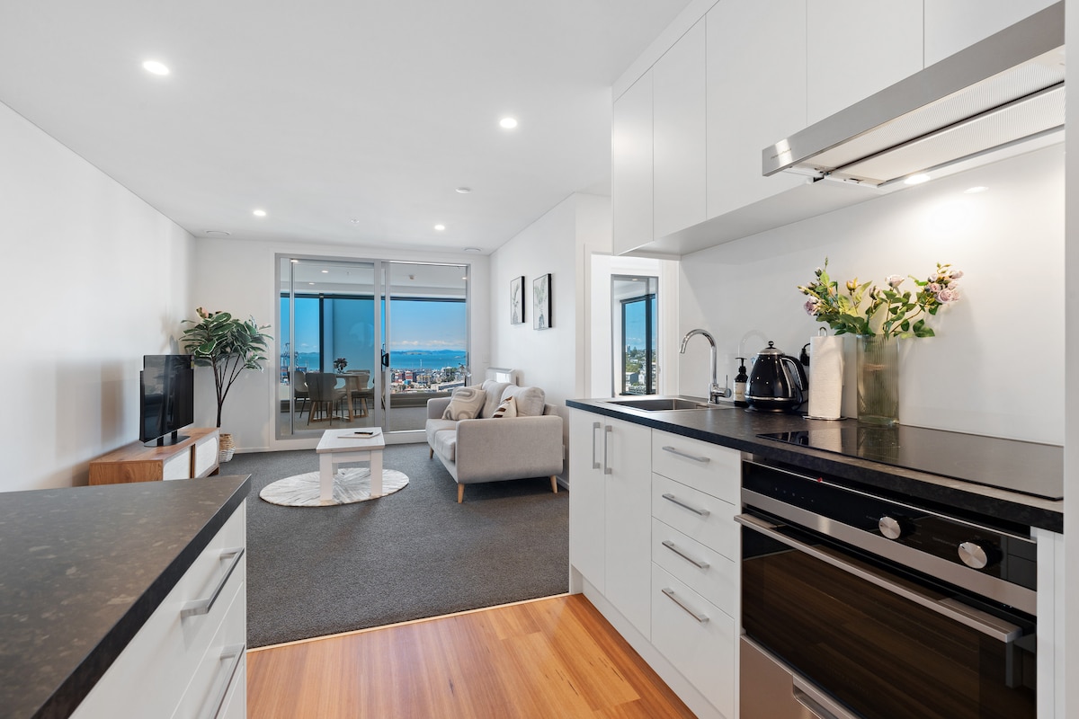 A modern kitchen area is visible, featuring sleek cabinetry, a stovetop, and an oven. Natural light floods the adjacent living space, where a comfortable sofa and a round rug are visible. Expansive windows frame a view of the sea and encourage a bright atmosphere.