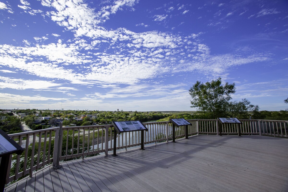 An observation deck is shown with wooden railings and a spacious floor. Informational displays are positioned along the edges. Below, the view extends over a river, with greenery and structures visible in the distance, under a sky filled with clouds and blue hues.