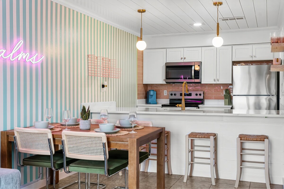 A modern dining area features a wooden table set for four, with light green chairs and tableware arranged on a pink placemat. Behind, a kitchen with white cabinetry displays a stainless steel refrigerator and a vibrant wall with pastel horizontal stripes. Soft lighting fixtures hang above.