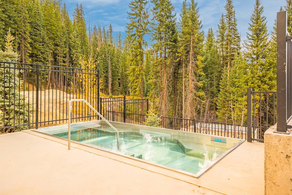 An infinity-style hot tub is situated on a concrete patio, surrounded by a black metal railing. Lush green trees create a natural backdrop, enhancing the serene outdoor setting. Water is gently bubbling in the tub, inviting relaxation in a secluded environment.