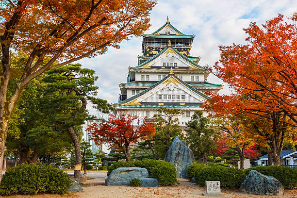 Osaka Castle is depicted amidst vibrant autumn foliage, showcasing its distinct architecture with a multi-tiered roof. The surrounding lush gardens and trees enhance the scene, while boulders and shrubs create an inviting natural setting.