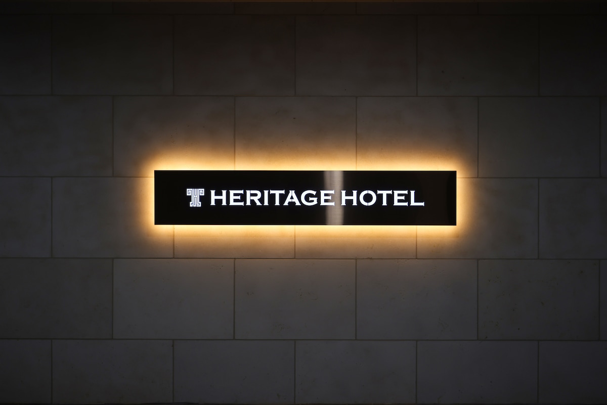 A sleek hotel sign displays the name 'Heritage Hotel,' illuminated by soft backlighting. The sign is mounted on a textured wall, creating a serene and contemporary appearance.