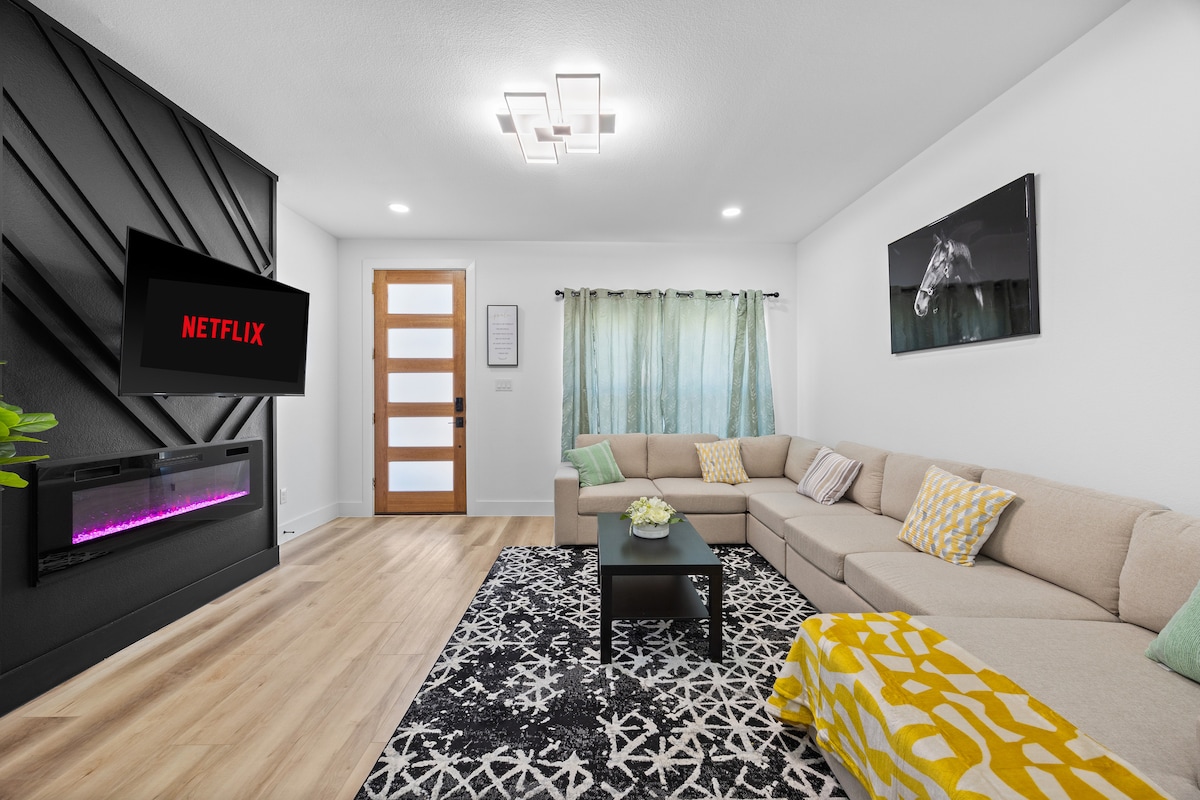 The living area features a modern sectional sofa with vibrant cushions arranged comfortably around a stylish area rug. A wall-mounted television presents a digital interface, while a sleek fireplace adds warmth. Large curtains frame the window, allowing natural light to filter into the space.