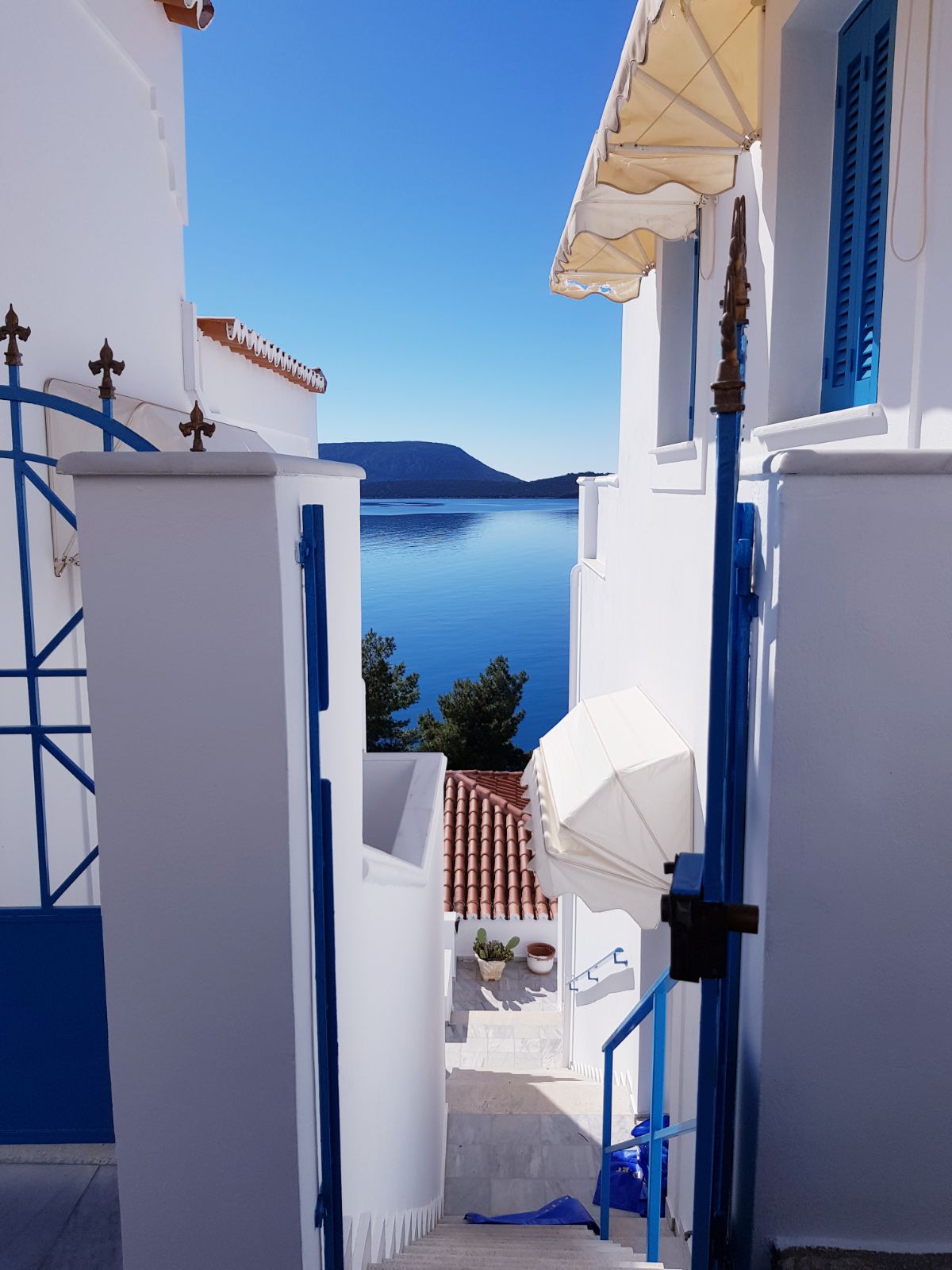 A view of the serene sea is framed by white walls and blue accents. Steps lead down towards the water, with a distant island visible across the calm surface. The bright blue sky enhances the tranquil atmosphere.
