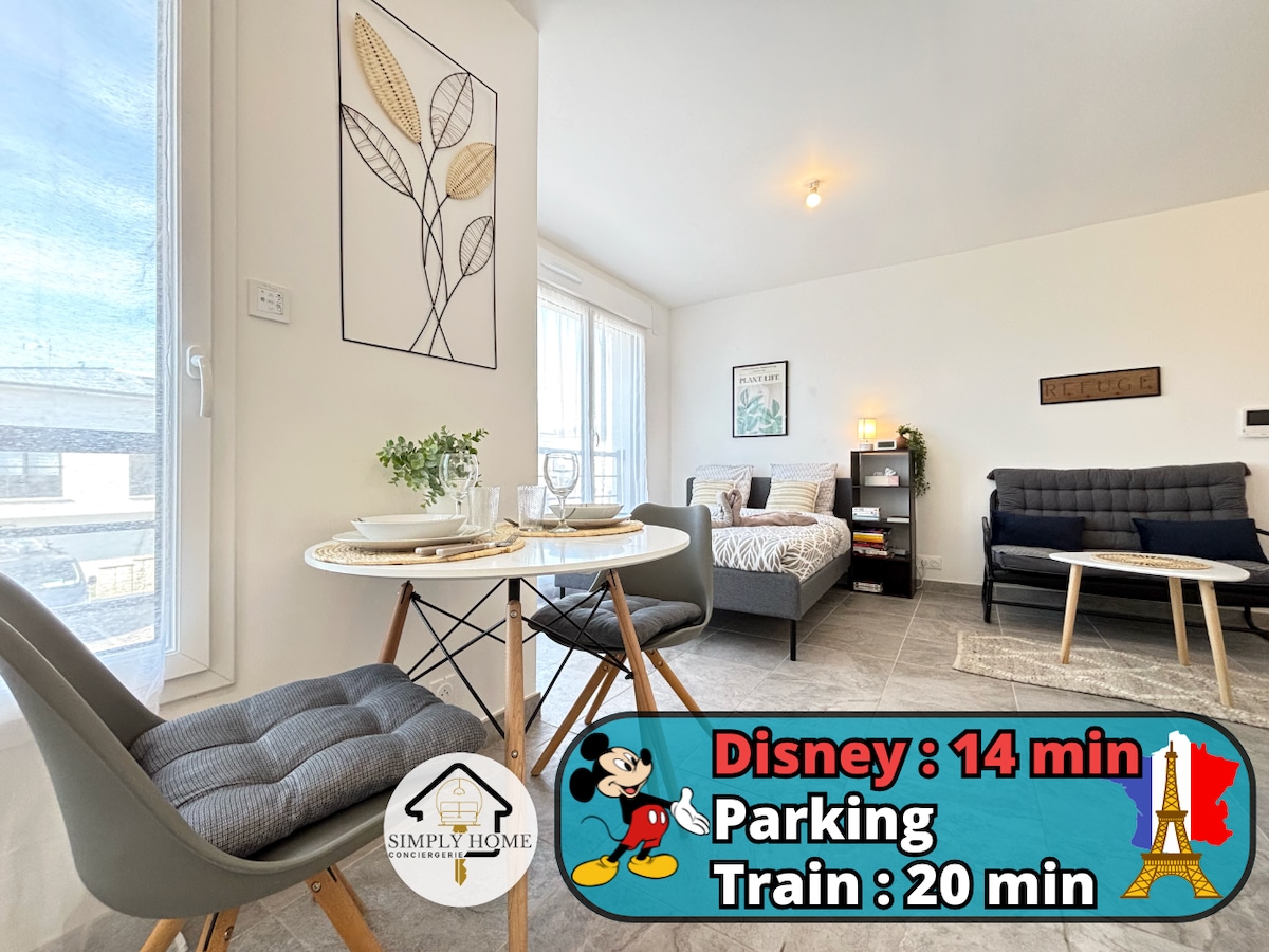 A bright living space features a comfortable bed and a well-arranged seating area. Natural light filters through large windows, creating a welcoming ambiance. A small dining table with two chairs is set near the window, complemented by decorative elements and a modern art piece on the wall.