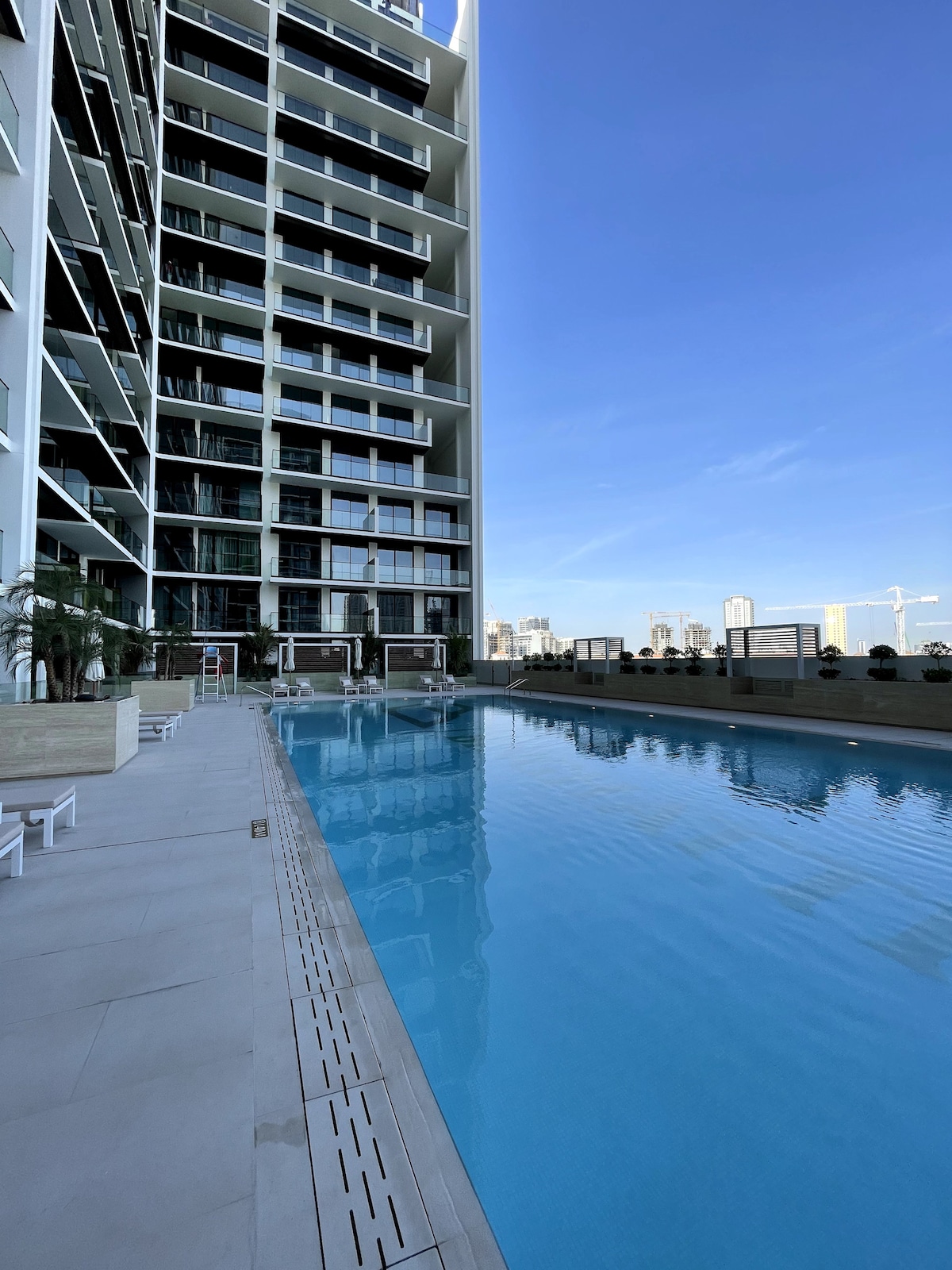 A modern swimming pool is framed by sleek architectural lines, with clear blue water reflecting the sky. Sun loungers are positioned alongside the pool area, and landscaped plants line the perimeter, creating a serene outdoor space for relaxation.