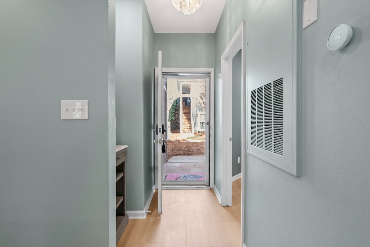 A welcoming entryway is enhanced by soft green walls and a wooden floor. A door opens to an outdoor space, allowing natural light to fill the area. A chandelier hangs from the ceiling, complementing the modern design elements.