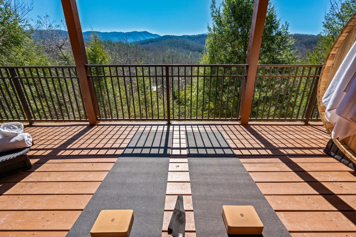 A serene outdoor space on the lower deck showcases two yoga mats positioned on a wooden surface. A barrel sauna and neatly arranged towels are visible to one side, with a backdrop of rolling mountains and clear blue skies enhancing the peaceful atmosphere.