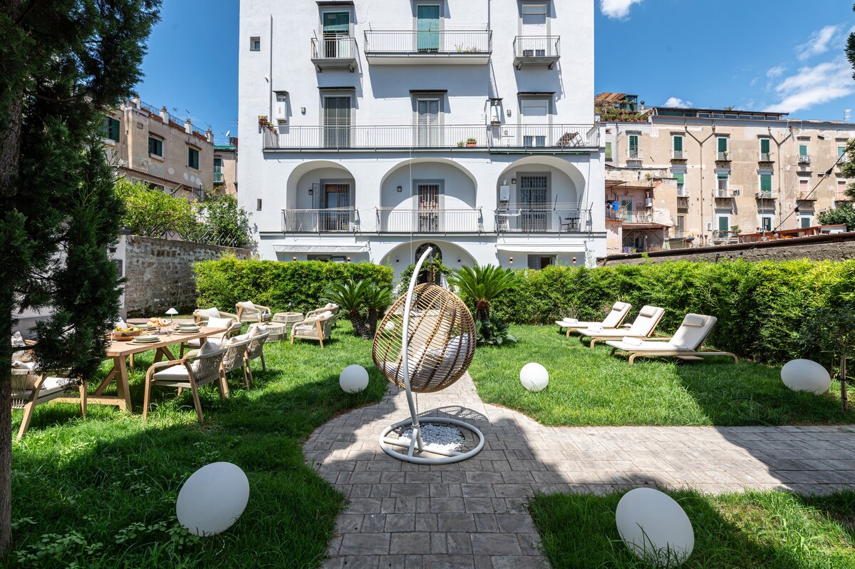 A spacious garden area is surrounded by greenery, featuring lounge chairs and a hanging seat. Dining tables are arranged on a grassy lawn, complemented by decorative orb lights. The backdrop showcases an apartment building with balconies and vibrant city structures beyond.