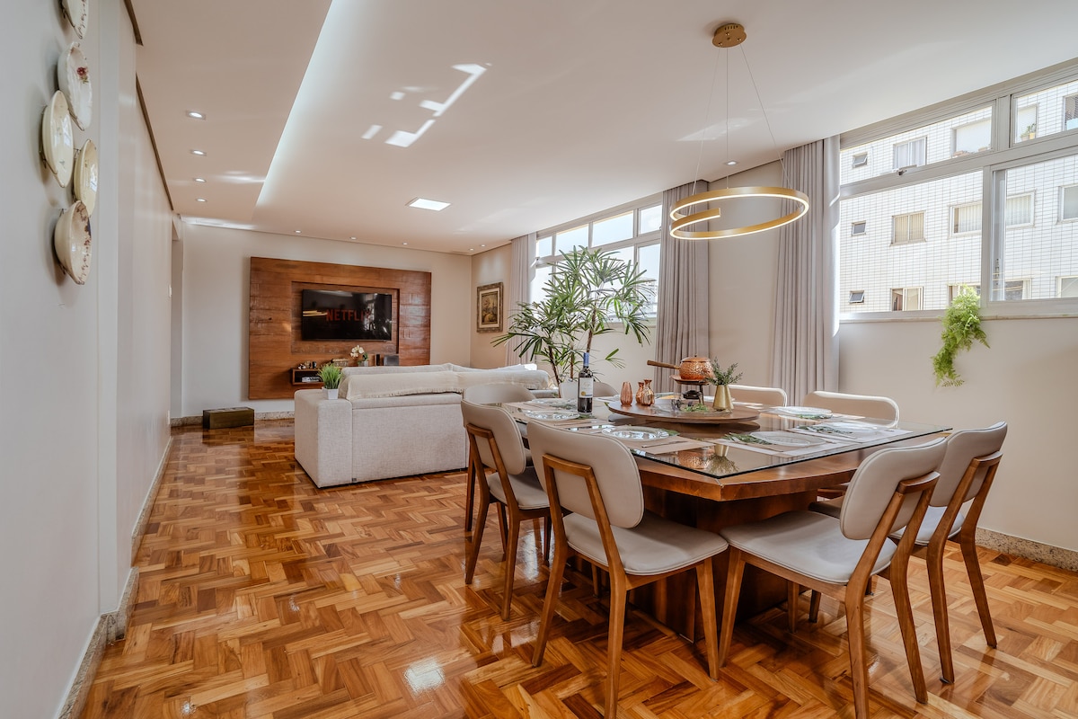 The living and dining area features light wood panel flooring and an elegant dining table set for eight. A comfortable sofa is positioned for relaxation, while natural light streams in through large windows adorned with soft curtains.