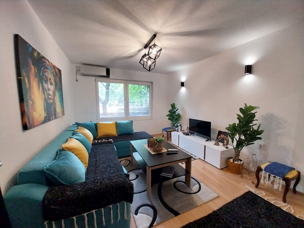 A well-lit living room features a large sectional sofa adorned with colorful cushions. A modern coffee table sits in the center, surrounded by decorative plants. A flat-screen TV is positioned on a white media console, while artwork and a cozy chair add character to the space.