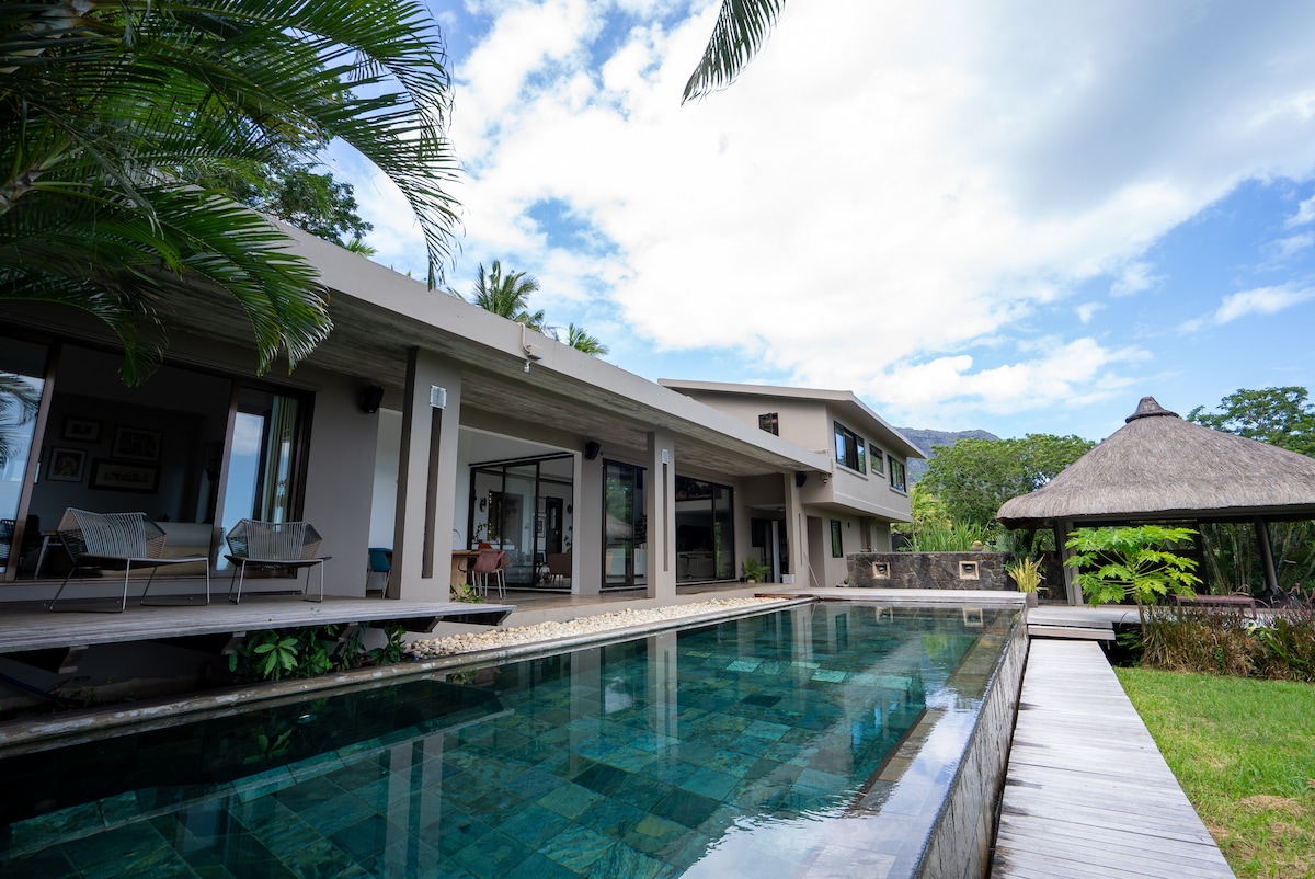 The villa exterior is highlighted by a serene infinity pool that reflects the sky, surrounded by a spacious wooden deck. Lush greenery and palm trees frame the area, while traditional thatched-roof structures add an elegant touch to the inviting outdoor space.