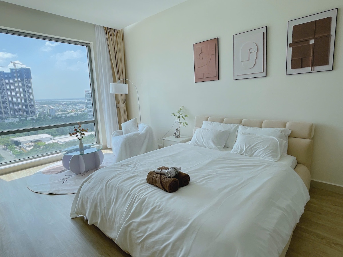 A spacious bedroom features a large, light-colored bed with crisp bedding and two pillows. Natural light floods the room through a large window, offering views of the surrounding area. A cozy seating area includes a soft chair and a small round table, complementing the serene atmosphere.
