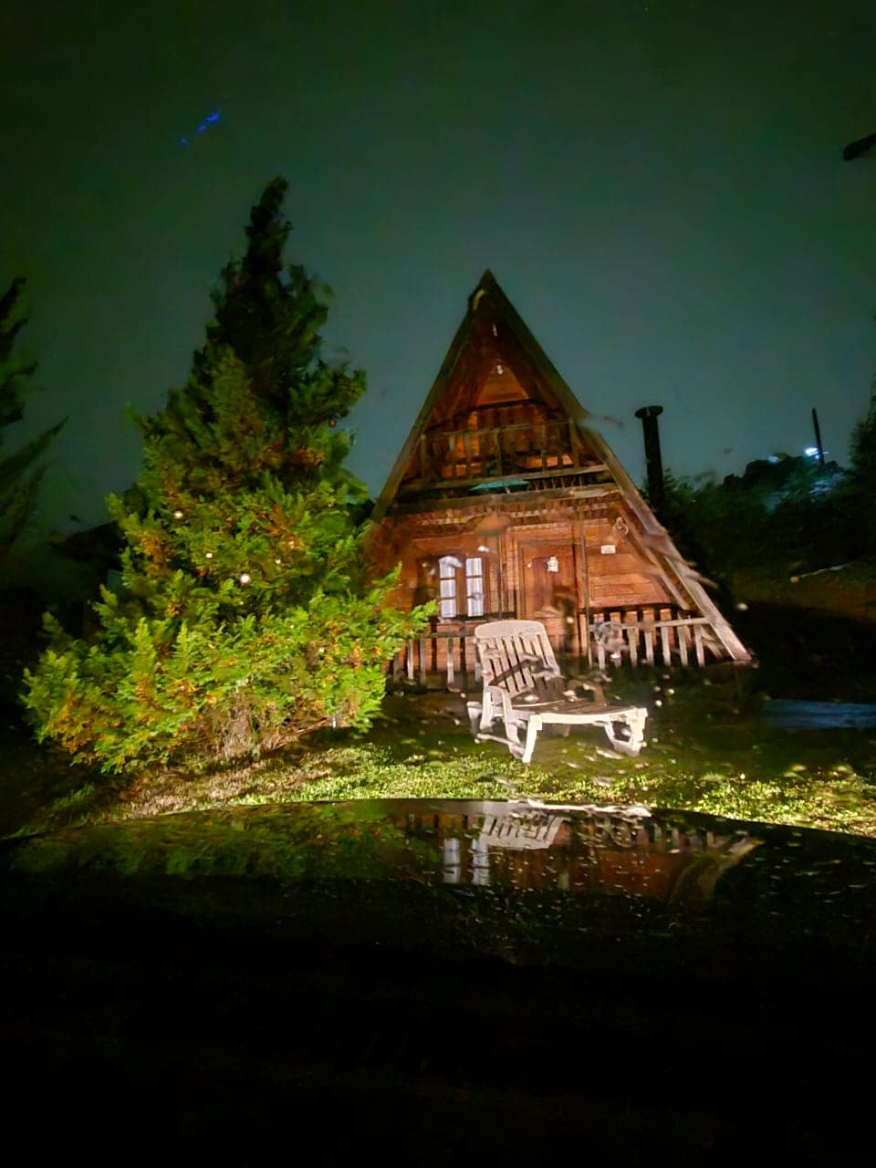 A charming wooden chalé is set under a dark sky, illuminated softly by surrounding lights. In front, a wooden chair is positioned beside a lush evergreen tree, with reflections visible in the water on the ground, creating a serene approach to the entrance.