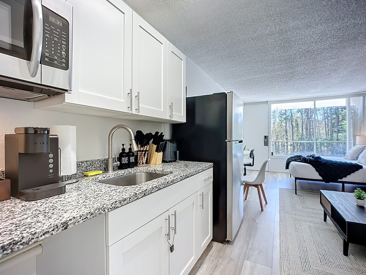 A modern kitchen area features sleek white cabinets and a granite countertop, with a microwave and coffee maker on display. A refrigerator is adjacent, and kitchen utensils are neatly arranged. A glimpse of the spacious living area with a bed and large windows is visible in the background.