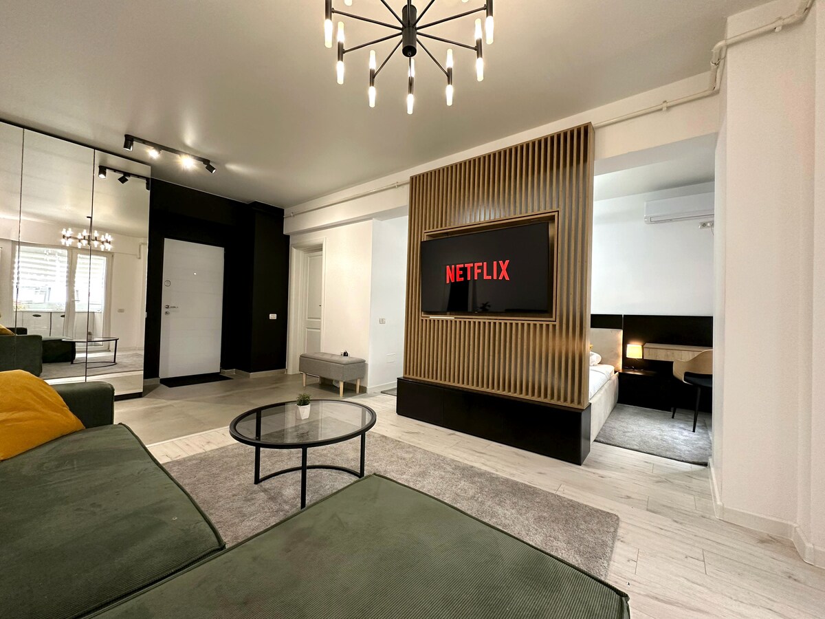 A modern living area features a comfortable dark green sofa and a round glass coffee table placed on a soft area rug. A large wall-mounted TV displays the Netflix logo, and a stylish wooden panel serves as an attractive backdrop. The space is well-lit, providing a welcoming environment.