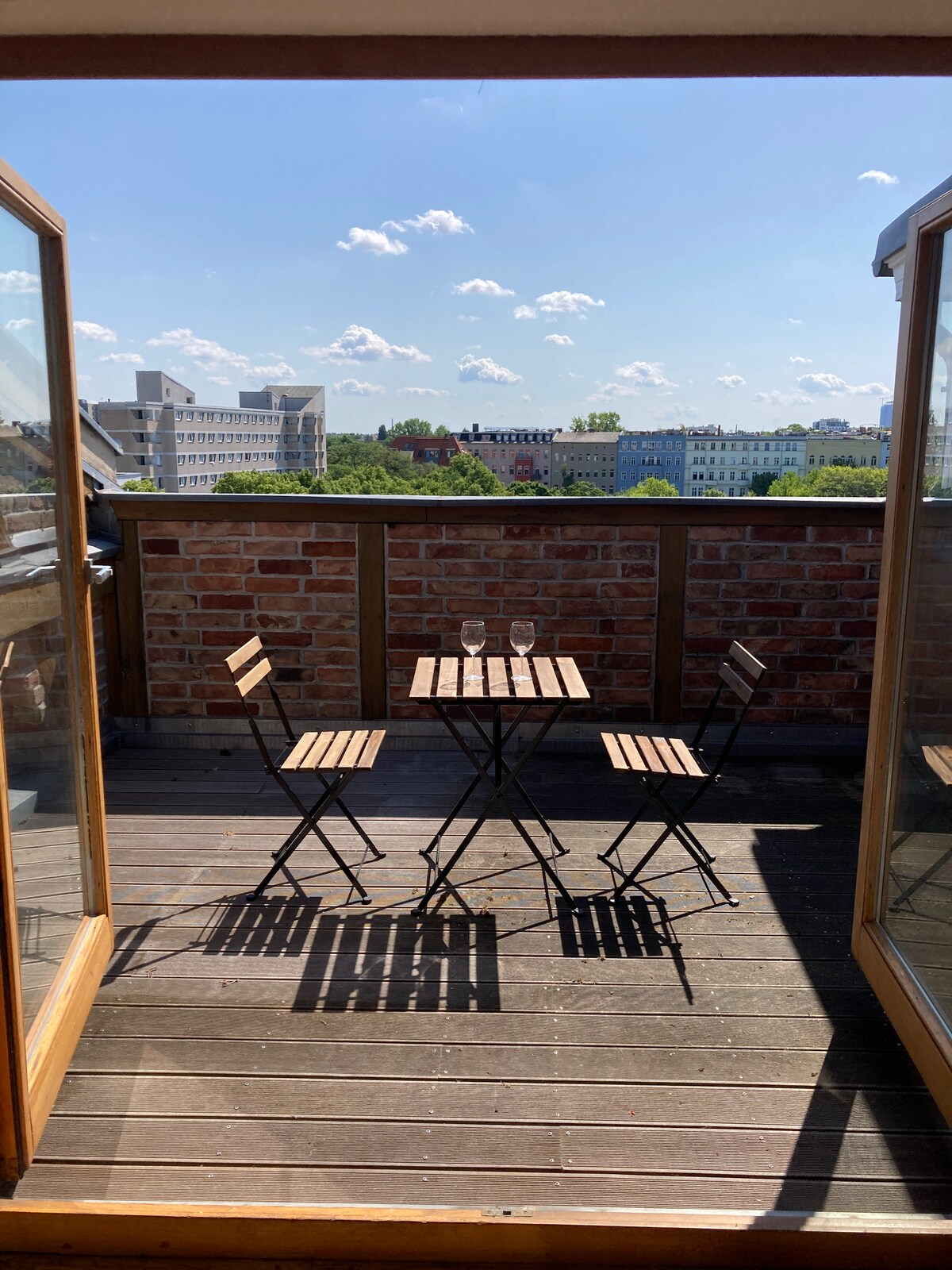 A private rooftop terrace features a small wooden table and two folding chairs, casting shadows on the wooden floor. The expansive city view is showcased against a clear blue sky, with distant buildings visible in the background.