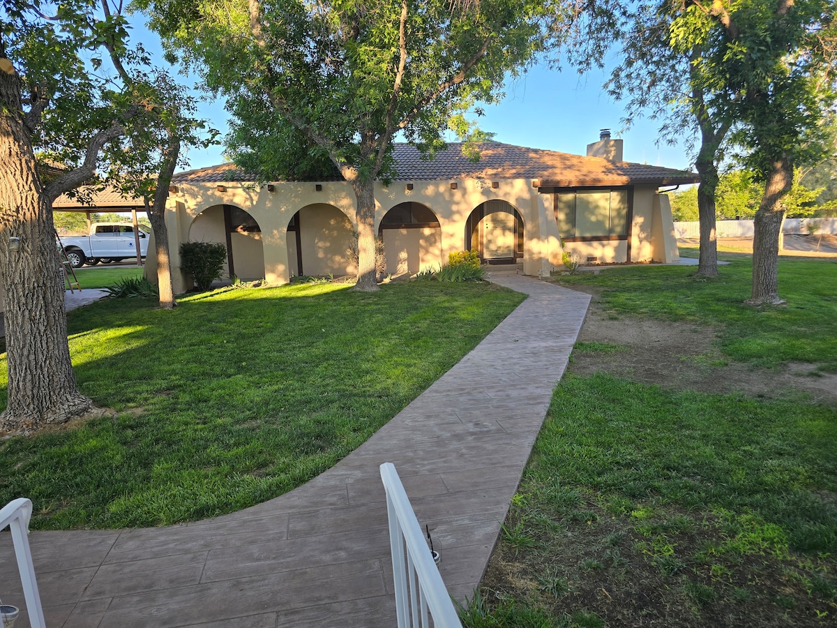 A serene exterior view of a single-story home set amidst lush green grass and mature trees. A winding pathway leads to the entrance, flanked by archways that enhance the building's architecture. Clear skies provide ample natural light to the surrounding area.