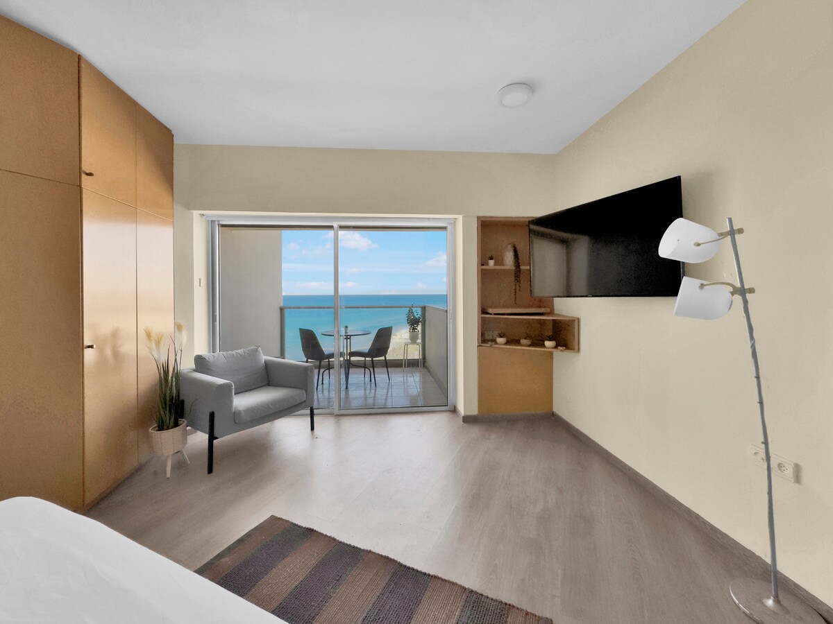 A bright, cozy retreat features a spacious room with light-colored walls and wooden flooring. A comfortable armchair is positioned next to a large sliding door that opens to a private balcony with sea views. A small outdoor table and chairs are visible on the balcony.