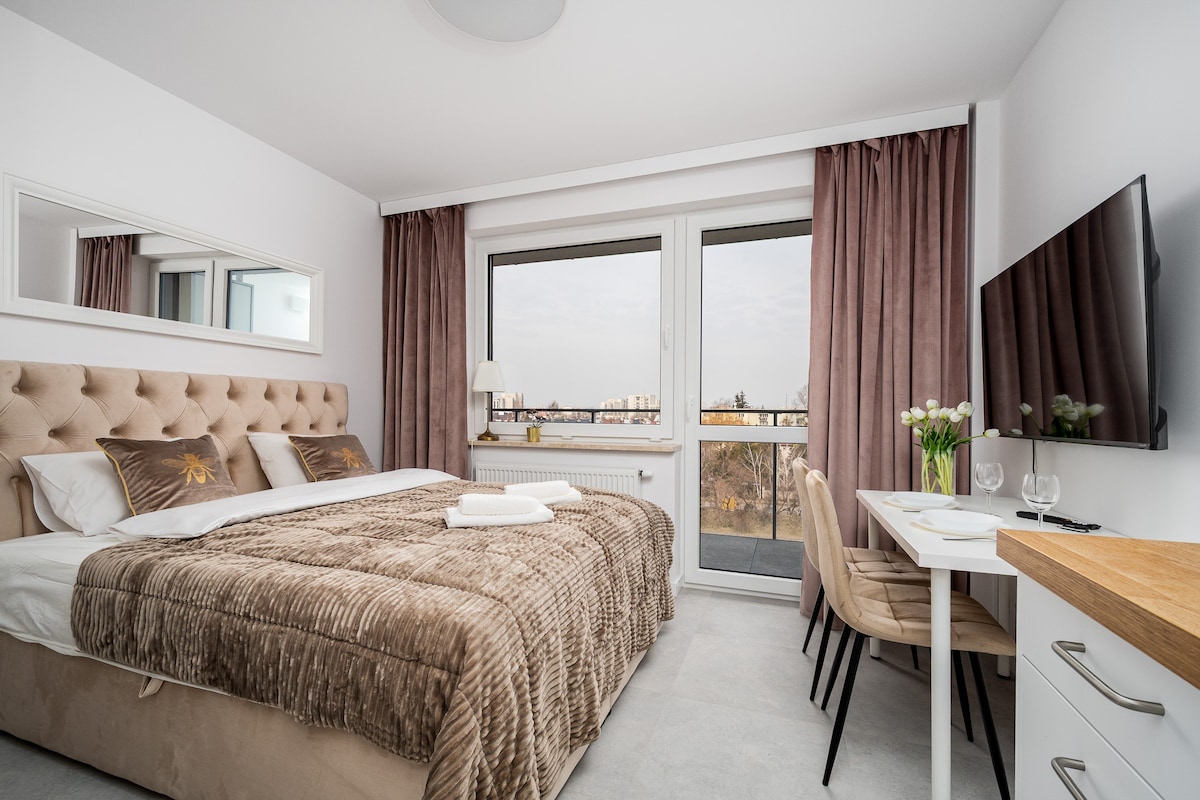 A bright bedroom features a spacious bed with a soft, textured blanket. Large windows offer views of the outside, while soft curtains frame the glass. A small dining table with chairs and a flat-screen TV complete the room's layout, enhancing the modern feel.