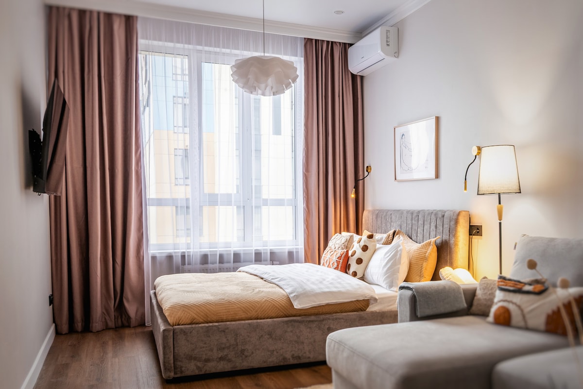 The bedroom features a spacious bed adorned with neutral-toned bedding and decorative pillows. Soft natural light enters through large windows dressed in sheer curtains. A cozy armchair and a television on the wall enhance the room's functionality.
