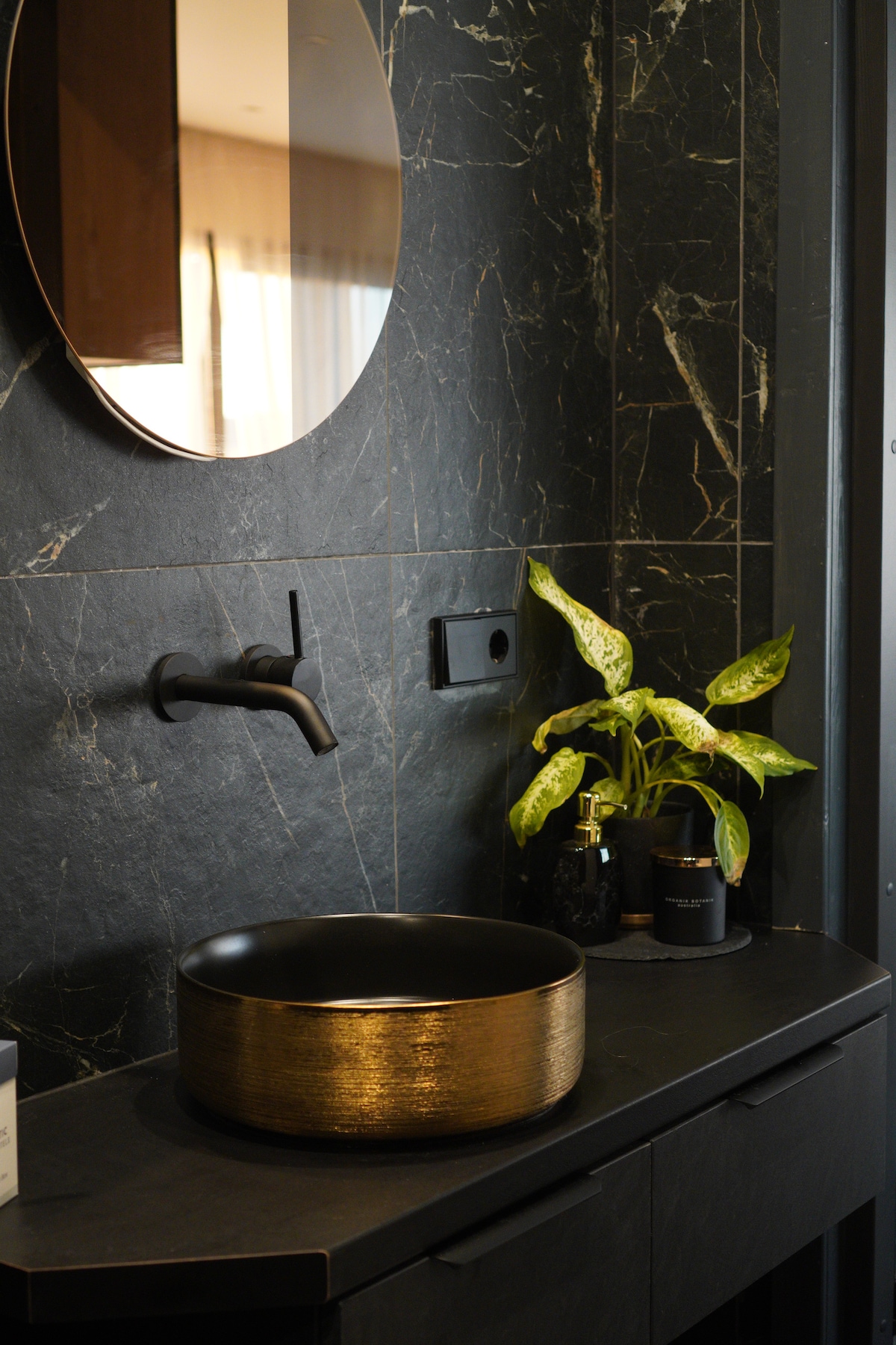 A modern bathroom is featured with a round mirror mounted above a dark stone countertop. A black and gold basin sits prominently, accompanied by a small potted plant and various toiletries. The sleek, textured wall adds a touch of elegance to the space.