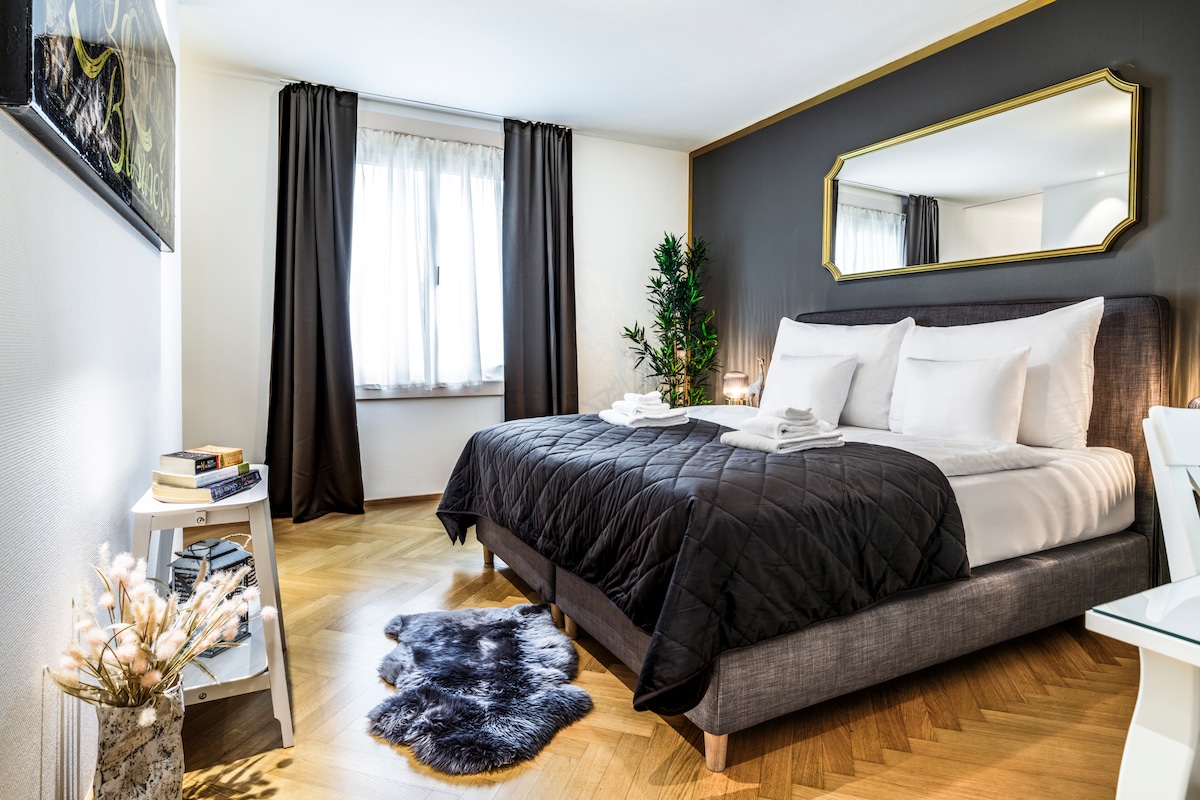 A spacious bedroom features a plush bed with crisp white linens and decorative pillows. Dark curtains frame the window, allowing soft light to enter. A stylish mirror reflects the room's elegance, while a small side table holds books and a decorative plant, adding a touch of greenery.
