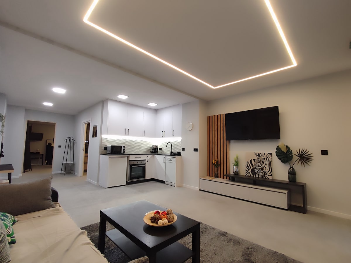 A modern living area features a comfortable sofa facing a low coffee table with a decorative bowl of fruit. A well-equipped kitchen is visible in the background, complemented by sleek cabinetry and a contemporary design. Soft lighting enhances the spacious ambiance.