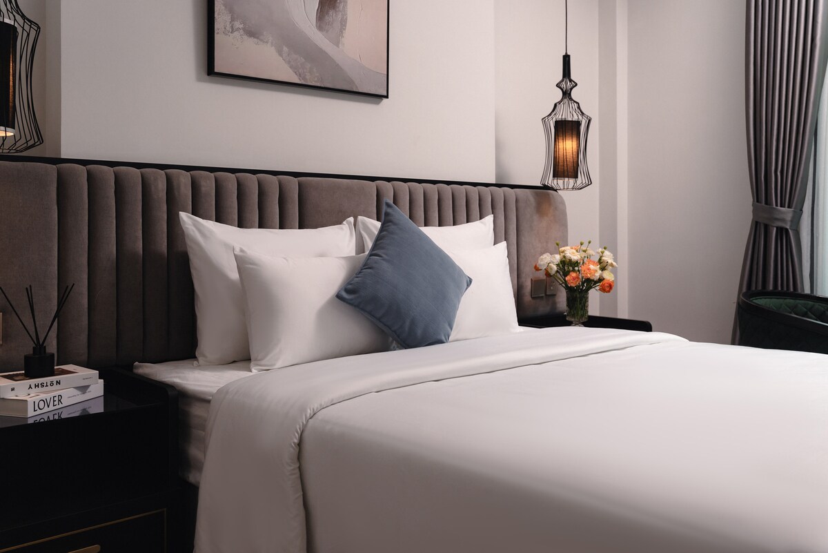 A neatly arranged sleeping area features a soft, spacious bed adorned with crisp white linens and a decorative blue pillow. Soft lighting is provided by pendant lamps on either side of the bed, while a vase of fresh flowers adds a touch of color.