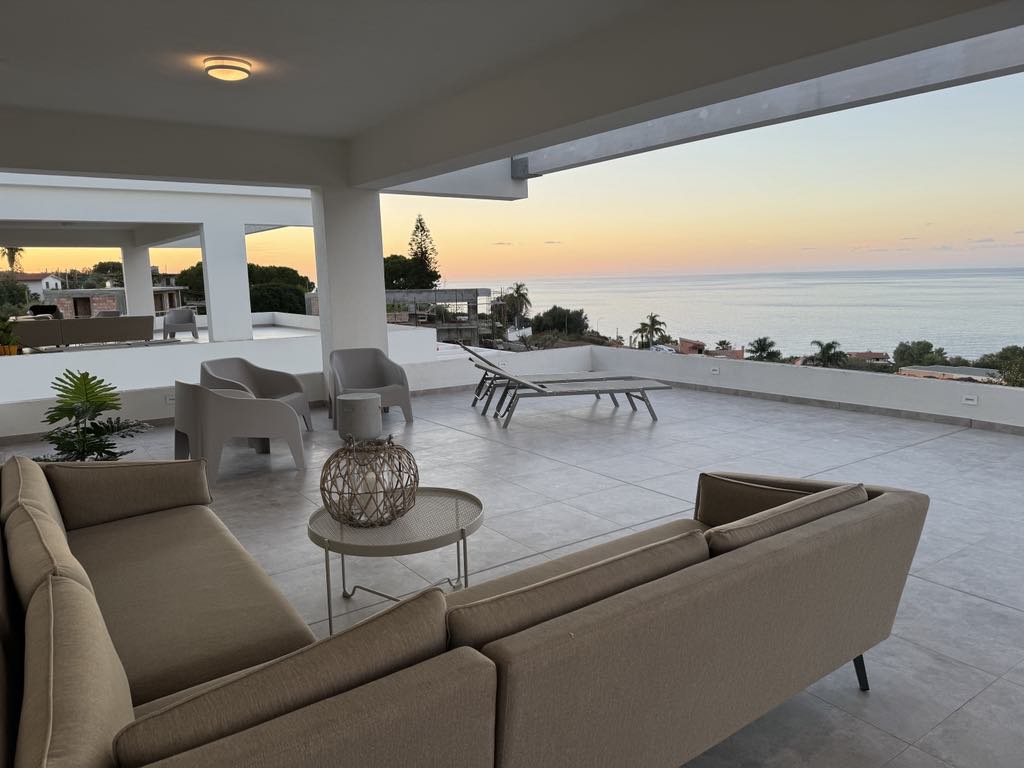 An inviting outdoor seating area features a comfortable beige sofa and light grey armchairs. A circular table is placed between the seats, and a sun lounger is positioned nearby. The view reveals a serene coastline with a pastel-hued sky at dusk.