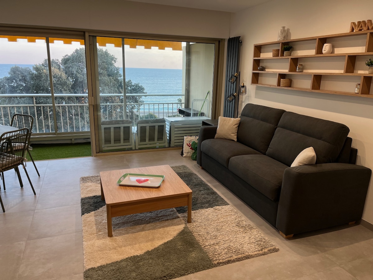 A cozy living area features a spacious dark sofa and a low wooden coffee table placed on a textured rug. Large glass doors provide a view of the sea and open to the terrace. Bookcases adorn the wall, complemented by a dining area with a table and chairs.